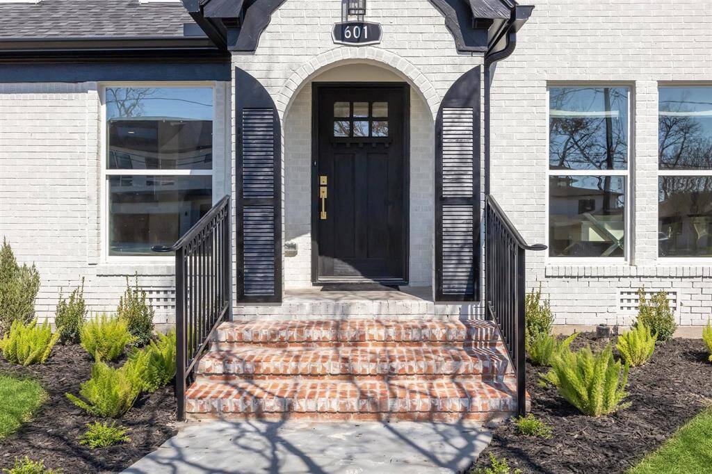 Charming entrance with a classic black front door and shutters, framed by white brick. The walkway features brick steps with iron railings, surrounded by well-maintained landscaping.