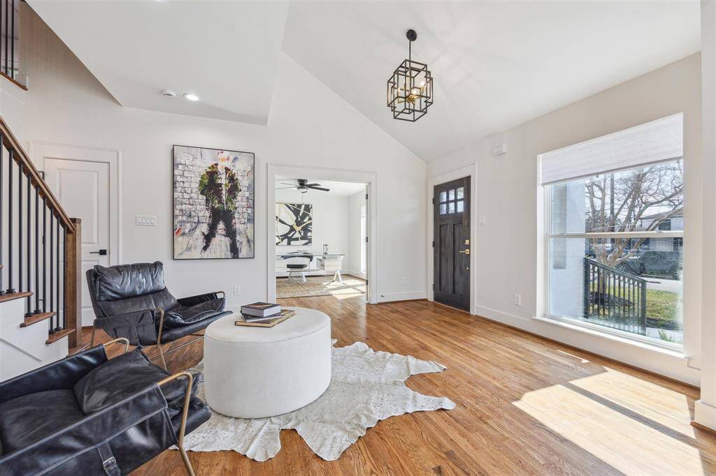 This bright and airy living room features hardwood floors, a modern light fixture, and large windows for ample natural light. The open layout includes a view into an adjacent room, while stylish seating and contemporary art add a chic touch.