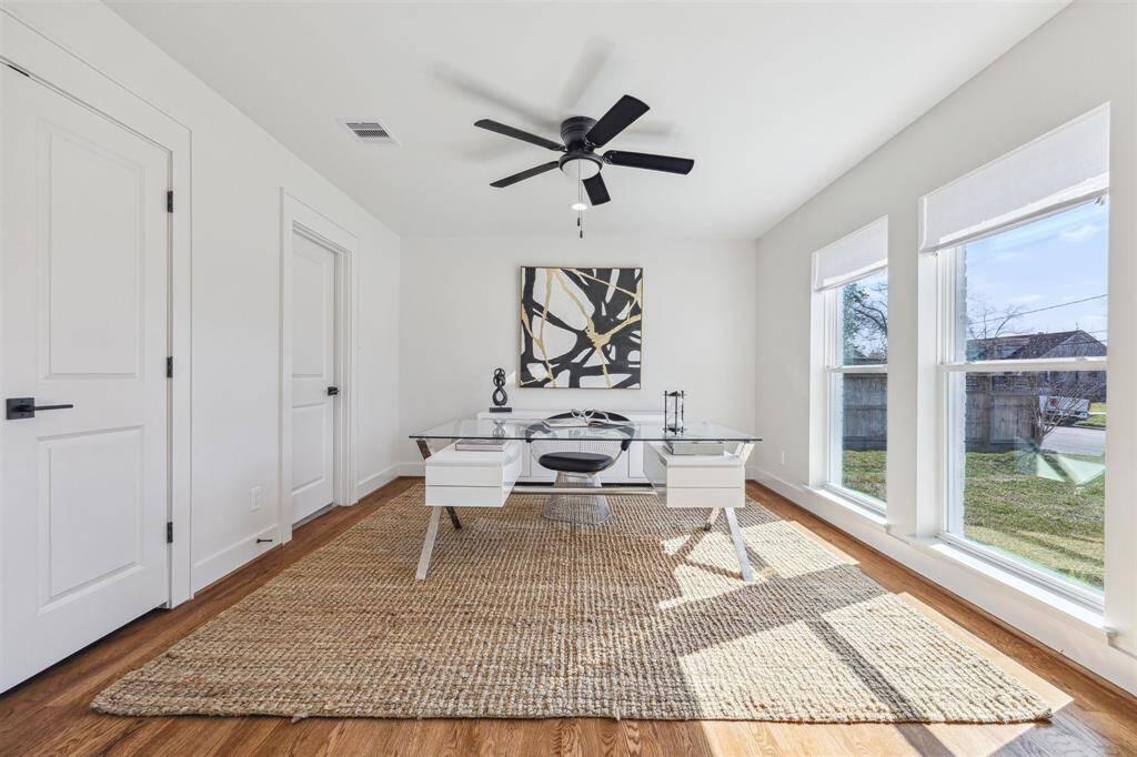 Bright and airy home office with large windows, a ceiling fan, and modern decor. Features a sleek glass desk and a natural fiber rug, providing a comfortable and stylish workspace.