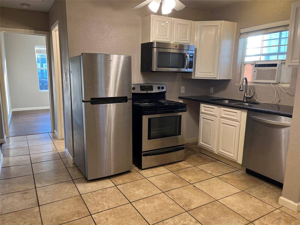 This kitchen features stainless steel appliances, including a refrigerator, oven, and dishwasher. It has beautiful cabinets, dark countertops, and tile flooring. An air conditioning unit is visible in the window, and there's a ceiling fan for added ventilation. The space is adjacent to one of the bedrooms.