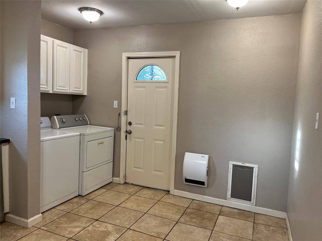 This photo shows a laundry space featuring a washer and dryer set, white cabinetry for storage, tiled flooring, and a door with a small window. The room has neutral walls and ceiling lights, creating a functional and bright space.