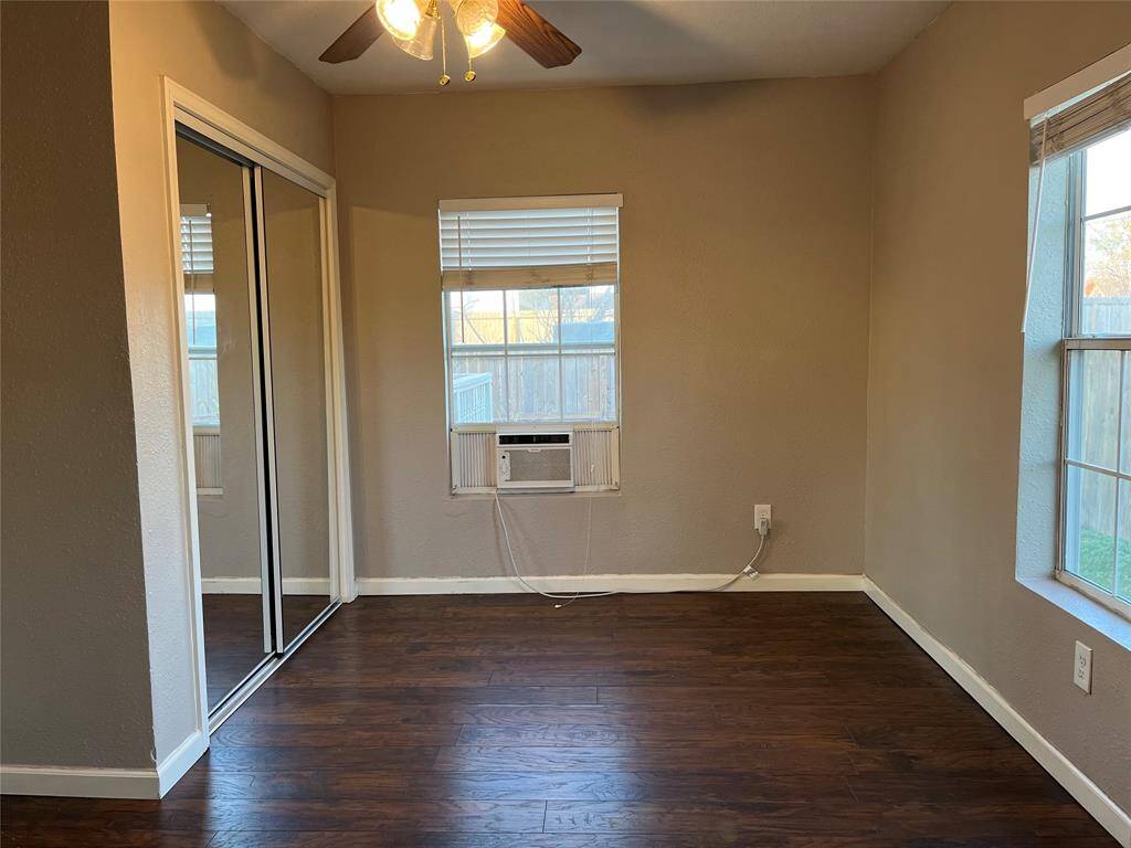 Cozy bedroom with dark floors, neutral walls, and ample natural light from two windows. It features a ceiling fan, mirrored closet doors, and a window air conditioning unit.