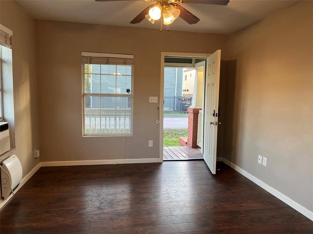 The living room features dark floors, neutral walls, a ceiling fan, and an AC unit. It has a window and a door leading to a small porch, providing natural light and easy outdoor access.