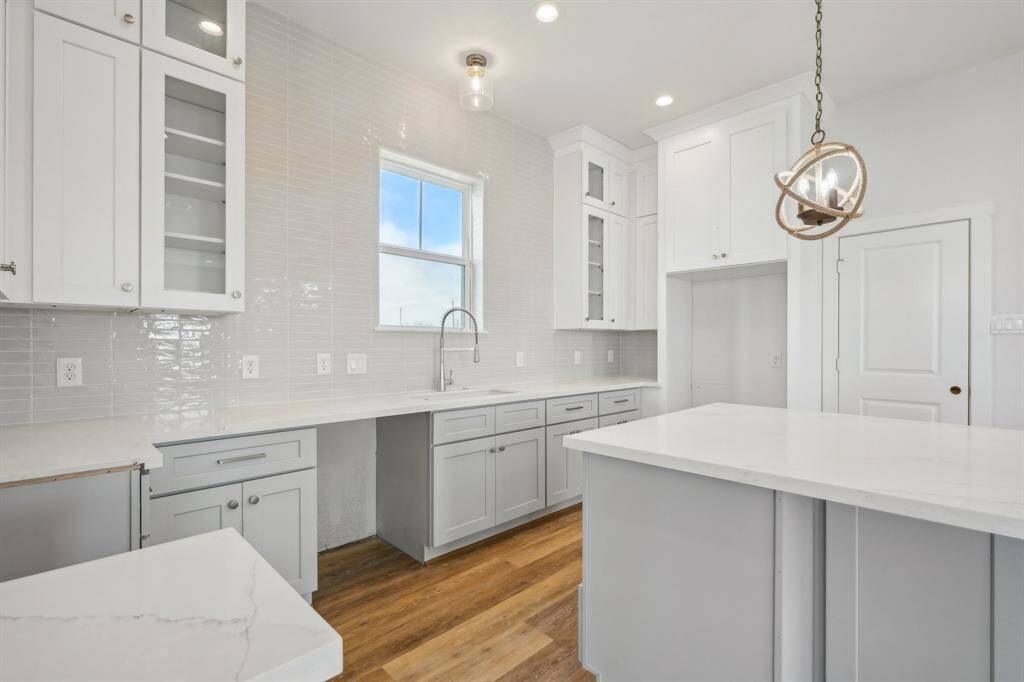 This bright, airy kitchen features soft gray shaker-style cabinets with glass-front doors, a sleek white quartz countertop, and a glossy, neutral-toned backsplash. The large window above the sink allows plenty of natural light to flood the space, highlighting the warm wood flooring.
