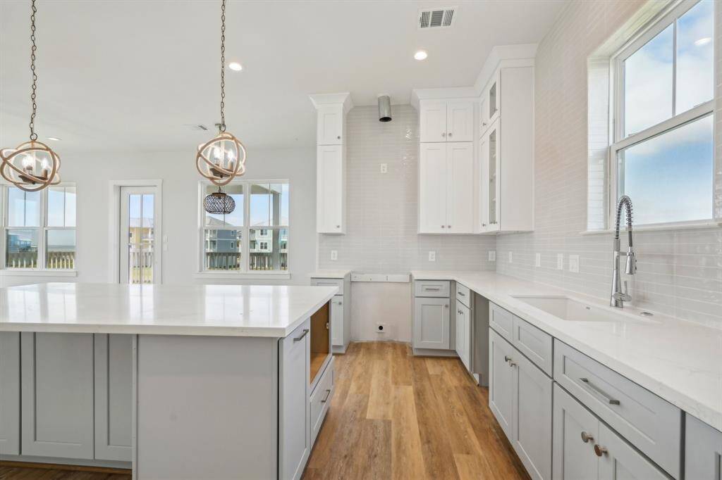 This bright, modern kitchen offers abundant storage with tall upper cabinets, glass-front doors, and a spacious island featuring extra cabinetry. White countertops, warm wood floors, and large windows complete the open, inviting design.