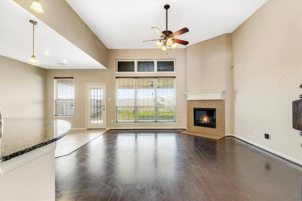 This spacious living room features high ceilings, large windows for natural light, and a cozy fireplace. It has an open floor plan with dark wood flooring and connects to a modern kitchen area with granite countertops. Ideal for comfortable living and entertaining.