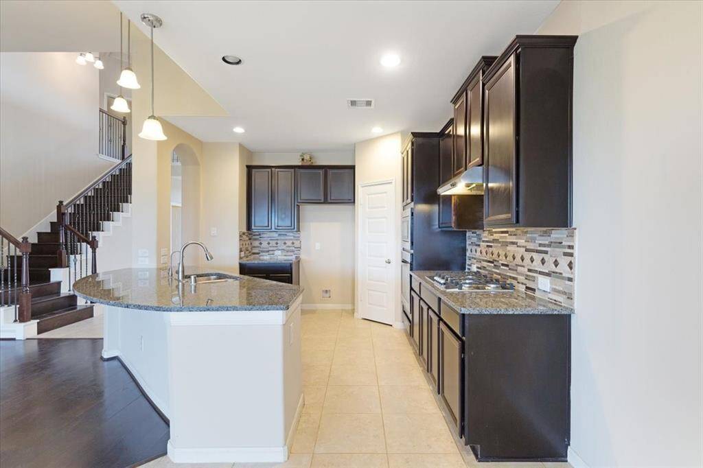 This spacious kitchen features dark cabinetry, granite countertops, and a stylish tile backsplash. It includes a modern island with a sink, pendant lighting, and an open layout leading to a staircase with elegant dark wood accents. Perfect for entertaining and family gatherings.
