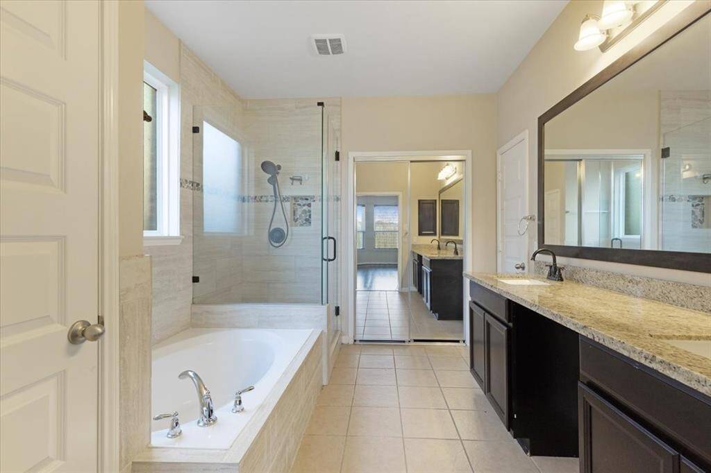 This bathroom features a spacious layout with a glass-enclosed shower, a separate bathtub, dual sinks with granite countertops, and ample storage in dark wood cabinetry. Natural light enhances the clean and modern design.