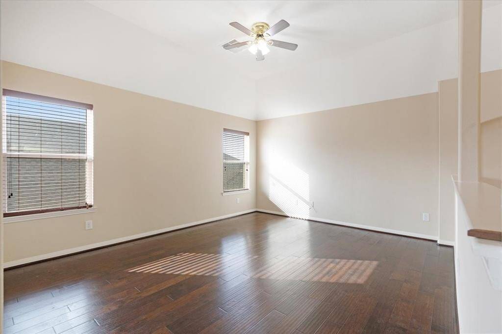 This spacious room features hardwood floors, neutral walls, and ample natural light from two large windows. A ceiling fan adds comfort, making it a versatile space for living or entertaining.