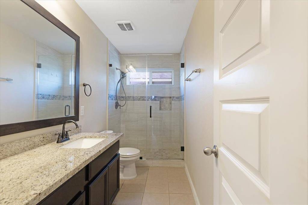 This is a modern bathroom featuring a granite countertop with a single sink, a large mirror, and a glass-enclosed shower. The space is well-lit with neutral tile flooring and wall colors, creating a clean and inviting atmosphere.