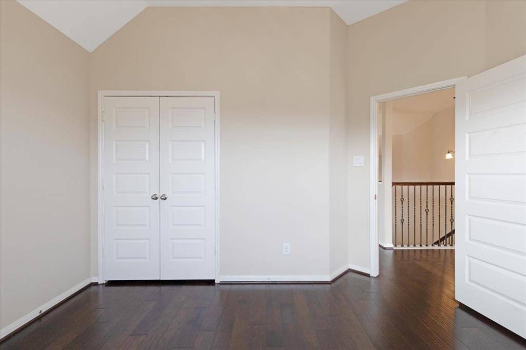 This room features neutral walls and dark hardwood flooring, with a double-door closet and a view of a staircase through an open doorway. It offers a clean and versatile space, ideal for a bedroom or office.