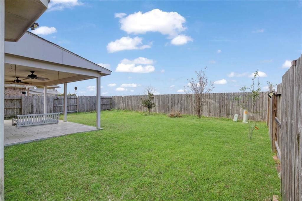 This photo showcases a spacious backyard with a well-maintained lawn, enclosed by a wooden fence. There's a covered patio area featuring a ceiling fan and a swing, ideal for relaxing or entertaining.