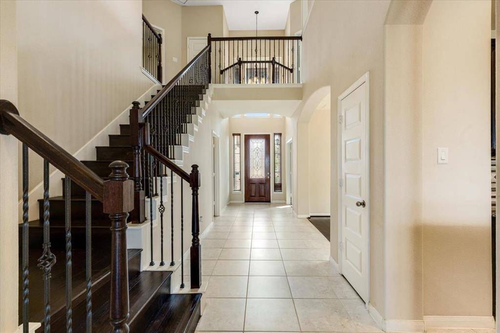 This photo showcases a spacious and elegant entryway with a high ceiling, featuring a grand staircase with dark wood and wrought iron railings. The tiled floor leads to a decorative front door with glass panels, providing a warm and inviting atmosphere.