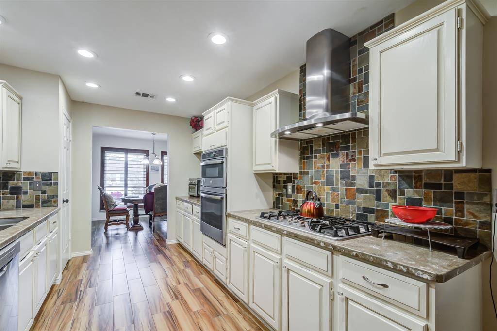 This kitchen features modern appliances, a gas stove with a sleek hood, and stylish multi-colored tile backsplash. It has ample cabinetry, wood look flooring, and connects to a bright dining area with large windows.