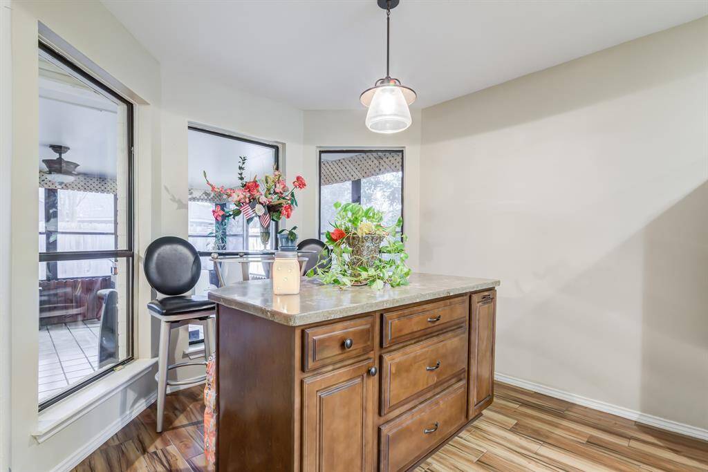 This bright kitchen area features a central island with wooden cabinetry and a stone countertop, illuminated by a hanging light fixture. Large windows provide ample natural light and offer views of an adjacent seating area.