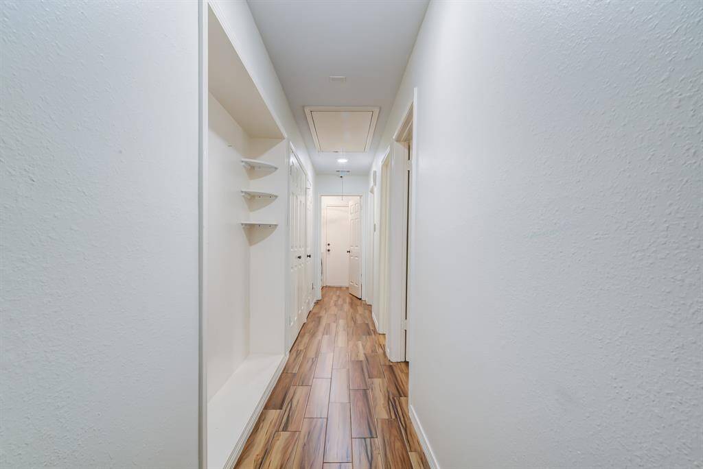 This photo shows a bright, narrow hallway with wood-look flooring and white walls. It features built-in shelves and multiple doors leading to different rooms.