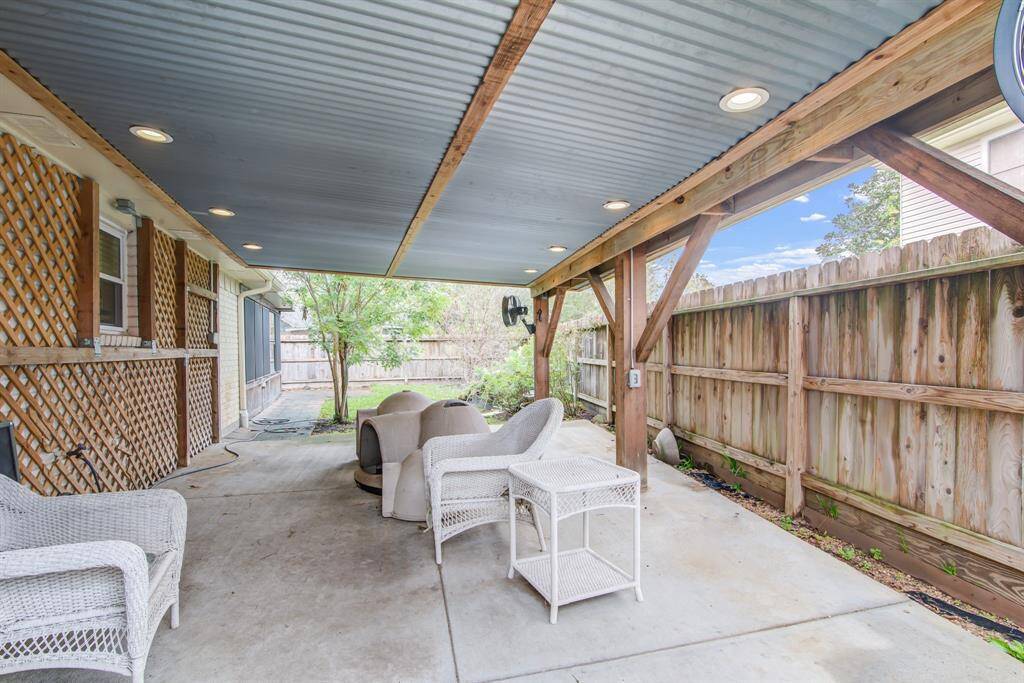 Covered patio with a metal roof, featuring cozy wicker seating and a fan. Surrounded by a wooden fence, it offers privacy and a view of greenery, perfect for outdoor relaxation.