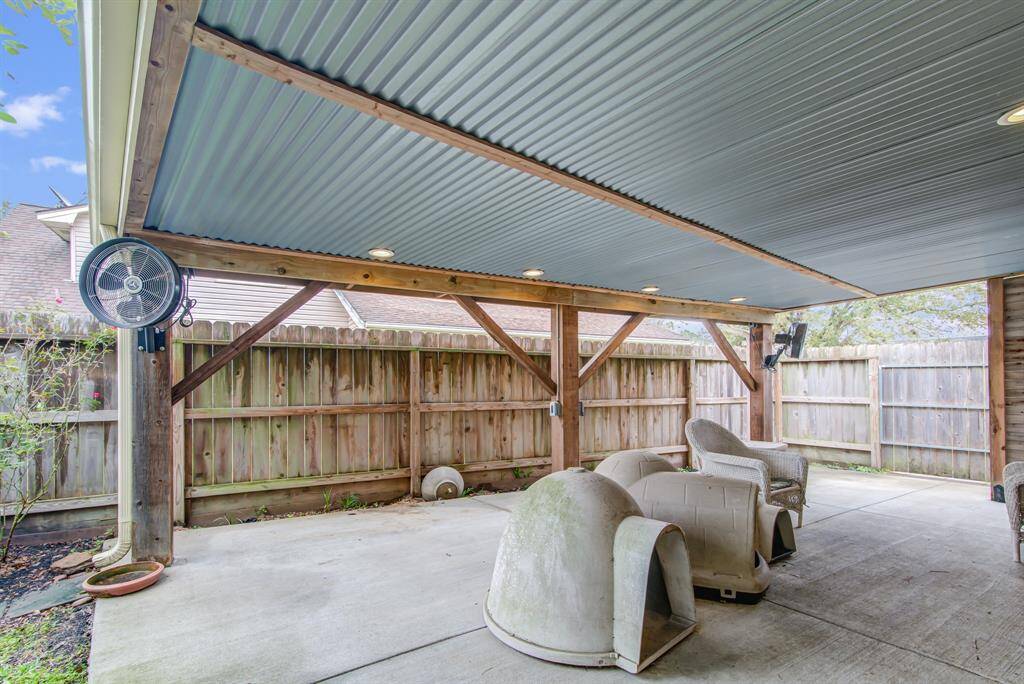 Covered patio with ample space, featuring a sturdy wooden frame and corrugated metal roof. Includes outdoor seating and fans, perfect for relaxation. Fenced for privacy.