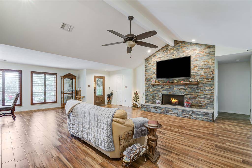 This spacious living room features a high ceiling with a ceiling fan, a cozy fireplace with a stone accent wall, and natural light from large windows. The open layout and wood-style flooring create a welcoming and stylish atmosphere.