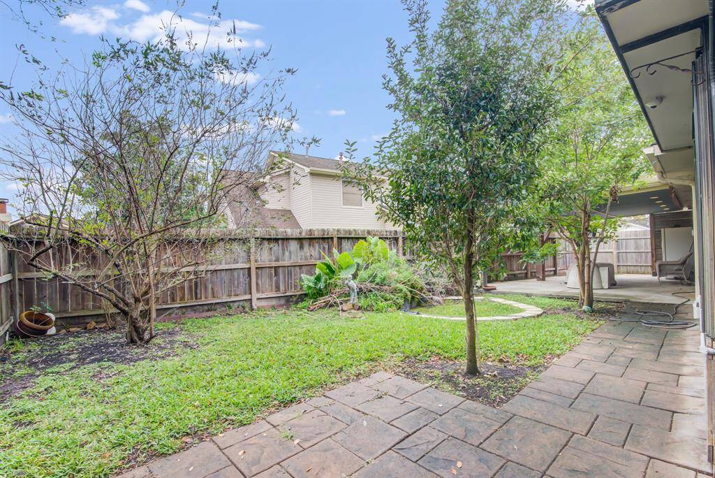 This backyard features a spacious patio with stone pavers, a well-maintained lawn, and mature trees providing shade and privacy. It's enclosed by a wooden fence, creating a cozy and private outdoor space ideal for relaxation or entertaining.