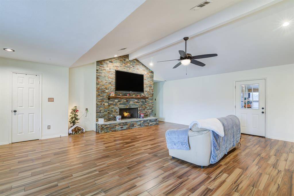 Spacious living room featuring a vaulted ceiling, modern wood-like flooring, and a cozy stone fireplace. The ceiling fan adds comfort, and the room is filled with natural light.
