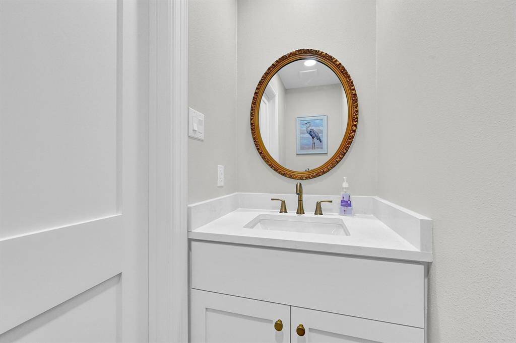 Charming half bath conveniently located near the entryway and formal dining area. Features a sleek white vanity with quartz countertop, elegant gold fixtures, and a decorative round mirror for a stylish touch. Perfect for guests and everyday convenience.