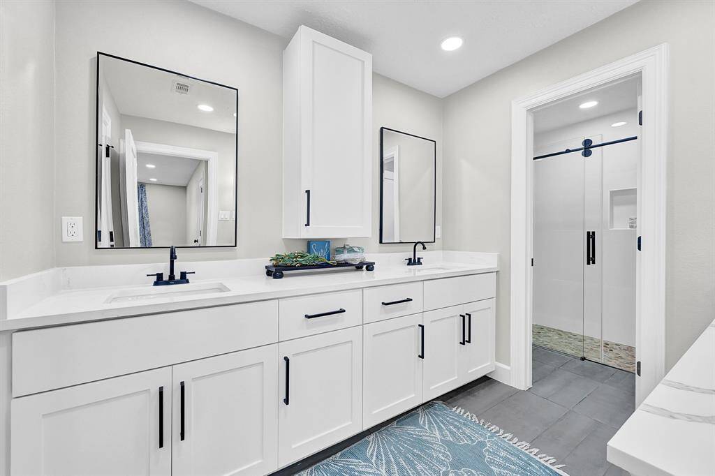 This luxurious master bathroom features a spacious dual-sink vanity with a built-in medicine cabinet, keeping toiletries, lotions, and shampoos neatly out of sight for a clutter-free look. The sleek quartz countertops, matte black fixtures, and single sliding frameless glass shower add a modern and elegant touch to this serene retreat.