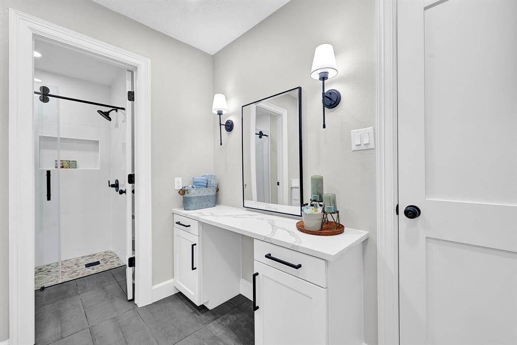 This luxurious master bathroom features a built-in makeup vanity with quartz countertops, perfect for a personalized beauty and grooming space. Elegant wall sconces, sleek cabinetry, and a frameless glass shower add to the modern charm and functionality of this serene retreat.