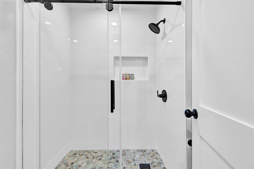 This luxurious master bathroom features a spacious walk-in shower with a sleek frameless glass door, complemented by elegant floor-to-ceiling tiles and a built-in shower niche for convenience. The pebble stone flooring and matte black fixtures add a spa-like touch to this modern retreat.