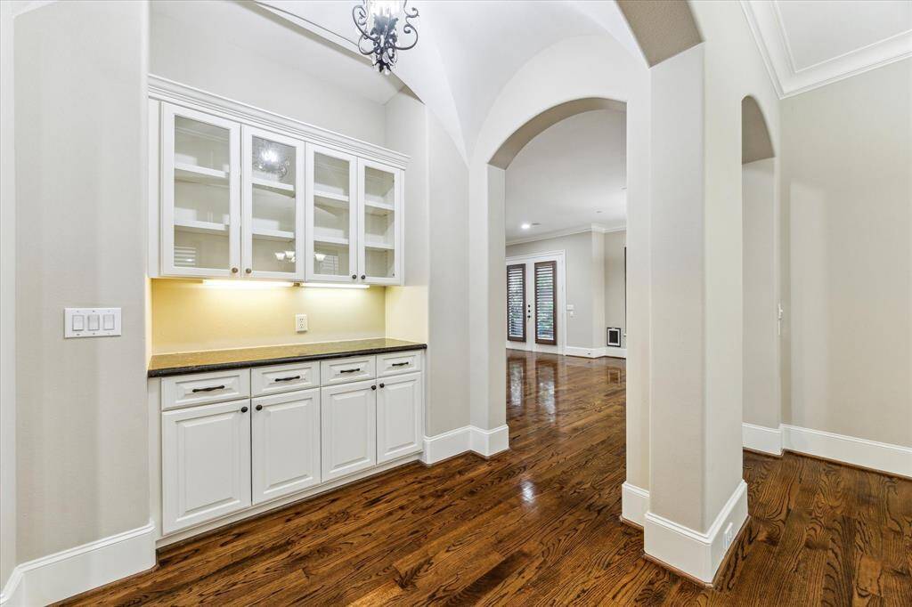 The elegant dry bar features a groin-vault ceiling with a wrought-iron pendant light, white cabinetry with glass-front upper doors, under-cabinet lighting, and a granite countertop. Perfectly positioned between the dining and family rooms, this sophisticated nook creates the perfect spot for happy hours or memorable dinner parties!
