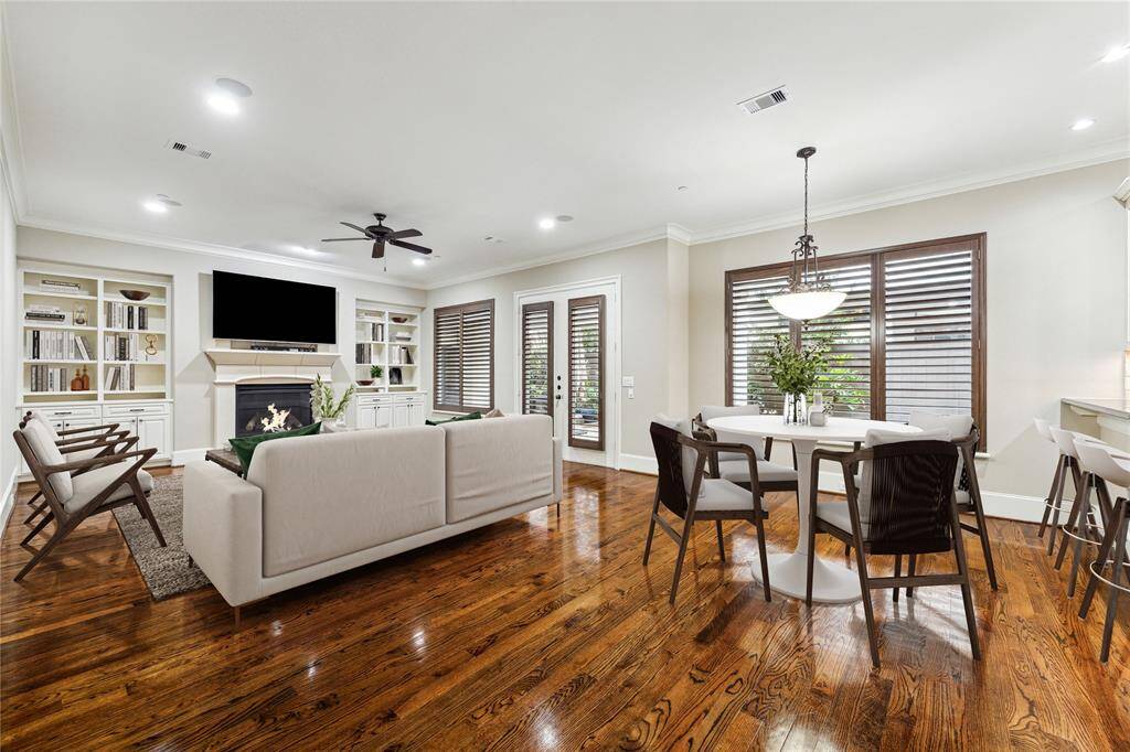 The open-concept family room features rich hardwood floors, a gas log fireplace with a stone surround flanked by built-in bookshelves with cabinetry, and an access door with plantation shutters to the back patio. Adjacent to this space, the breakfast room is highlighted by a decorative wrought-iron pendant light and offers a bright and airy setting for casual dining. This photo has been virtually staged.