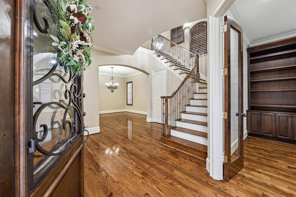 Welcome guests through a solid wood front door with intricate wrought iron and glass detailing into the grand foyer, where rich hardwood floors and soaring 9’11” ceilings create an inviting first impression. A stately staircase with wood treads and decorative iron railings leads to the second floor, while an arched opening leads to the dining room and double doors open to the private study.