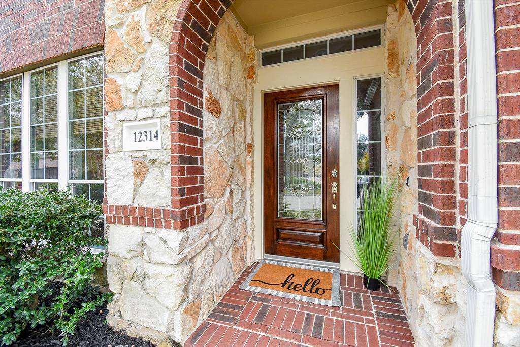 The front entrance welcomes you with a stately arched doorway and a warm, decorative glass door. The walkway, accented with brick details, leads to a picturesque front yard framed by vibrant flower beds and lush greenery, setting the perfect tone for this residence.