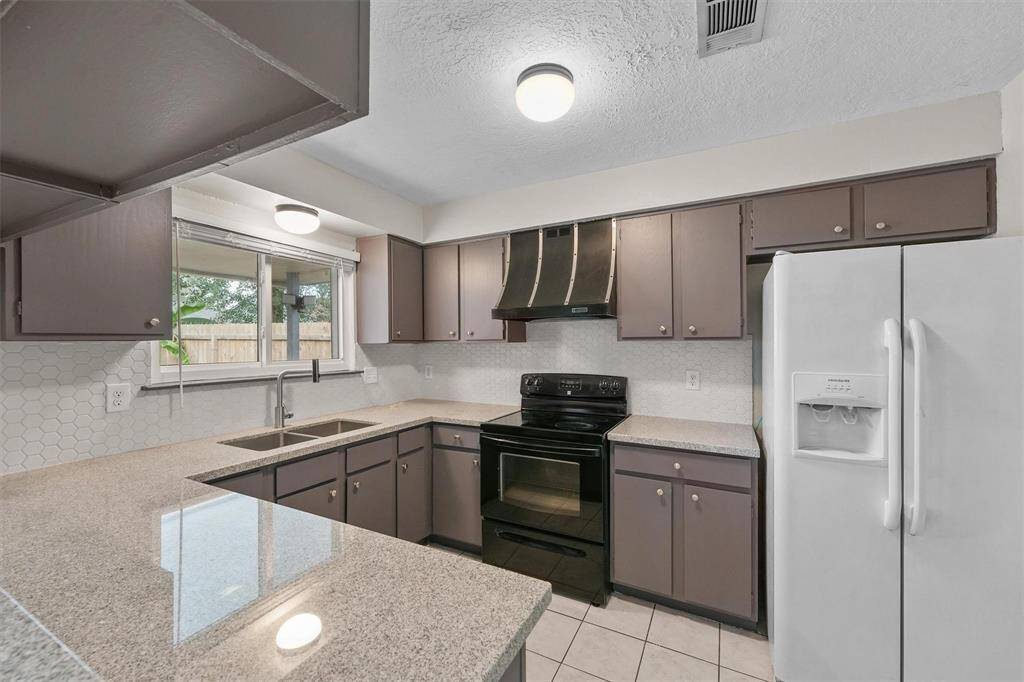 This kitchen features modern appliances, ample countertop space, and neutral cabinetry. It includes a double sink under a bright window, a sleek black stove with an overhead hood, and a double-door refrigerator. The design offers a practical and inviting space for cooking and entertaining.
