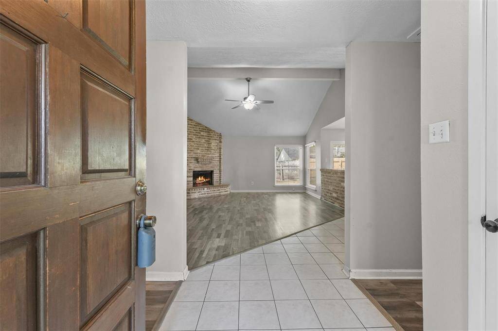 This inviting entryway features a warm, wooden door that opens into a bright living area with light tile flooring transitioning to wood. A cozy brick fireplace and large windows enhance the space, creating a welcoming atmosphere.