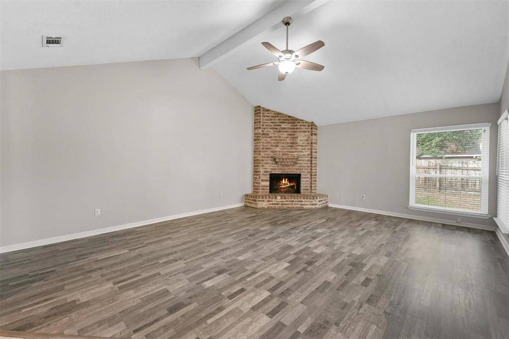 This spacious living room features a vaulted ceiling with a ceiling fan, a cozy brick fireplace, and large windows that provide plenty of natural light. The neutral walls and wood-look flooring offer a modern and inviting atmosphere.