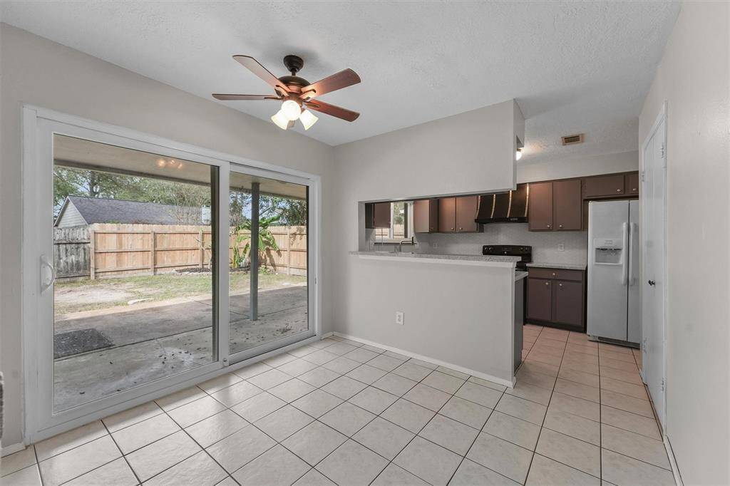 Bright dining area with tiled flooring, a ceiling fan, and large sliding glass doors leading to a fenced backyard. Adjacent is a kitchen with dark cabinetry, a sleek countertop, and modern appliances.