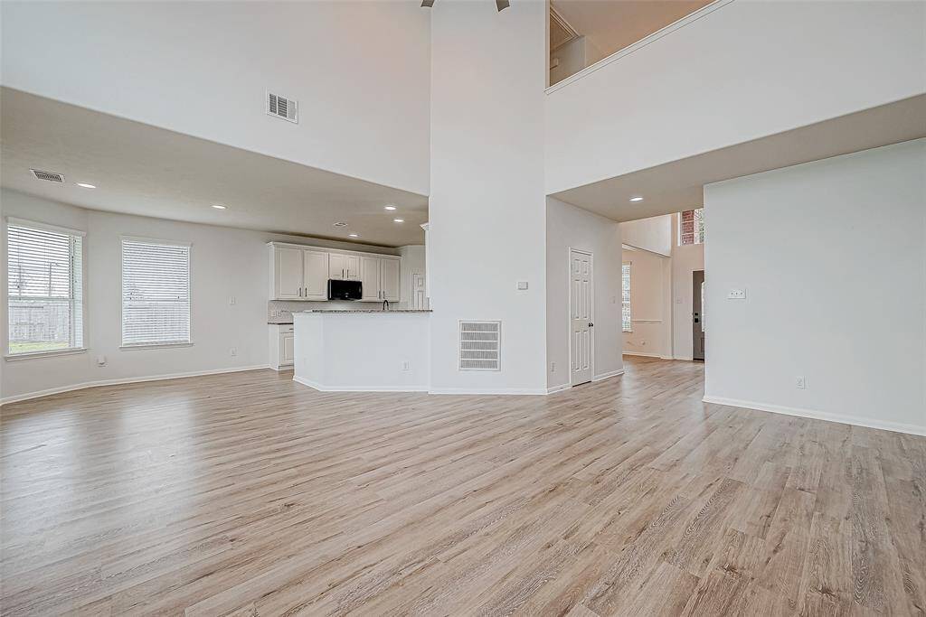 This spacious open-concept living area features high ceilings, abundant natural light, and wood-look flooring. The modern kitchen includes white cabinetry and a breakfast bar, enhancing the home's inviting and airy atmosphere.