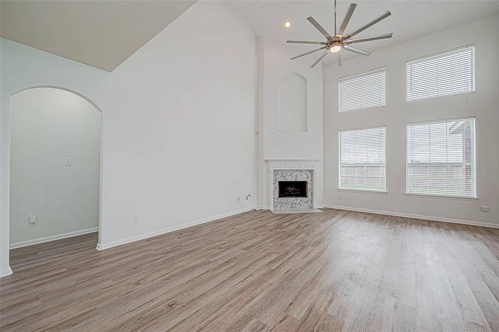 Bright and spacious living room with high ceilings, large windows, and a modern ceiling fan. Features a cozy fireplace with a stylish tile surround and light wood flooring throughout.