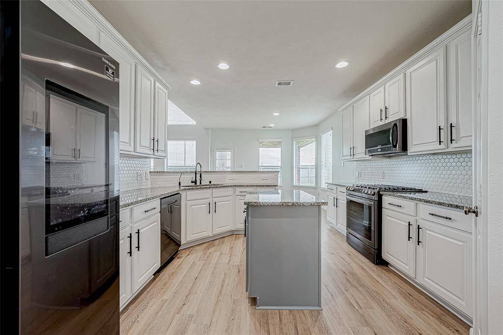 This bright, modern kitchen features white cabinetry, stainless steel appliances, and a central island with granite countertops. The space is enhanced by natural light from multiple windows, complemented by wood flooring and a stylish hexagonal tile backsplash.