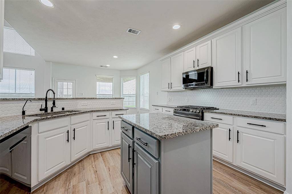 Modern kitchen with granite countertops, white cabinetry, stainless steel appliances, and a central island. The space is bright and airy with ample natural light and wood flooring.