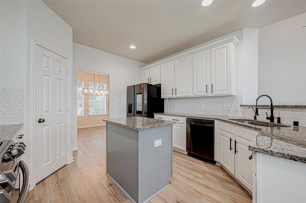 Modern kitchen with granite countertops, white cabinetry, and stainless steel appliances. It features a center island and hardwood floors, with ample natural light from an adjacent dining area.