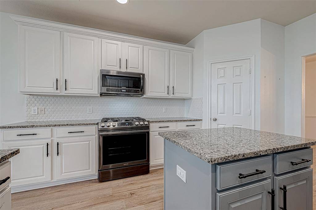 This kitchen features sleek granite countertops, white cabinetry, a central island with ample storage, and modern appliances, including a built-in microwave and stainless steel oven. The light wood flooring and hexagonal tile backsplash add a contemporary touch.