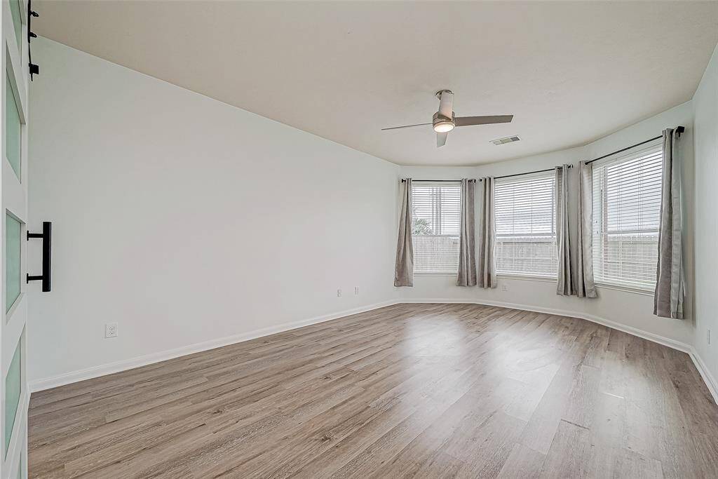 The primary bedroom with large bay windows, neutral walls, and light wood flooring. Features a modern ceiling fan and double doors. Ideal for a cozy living area or bedroom.