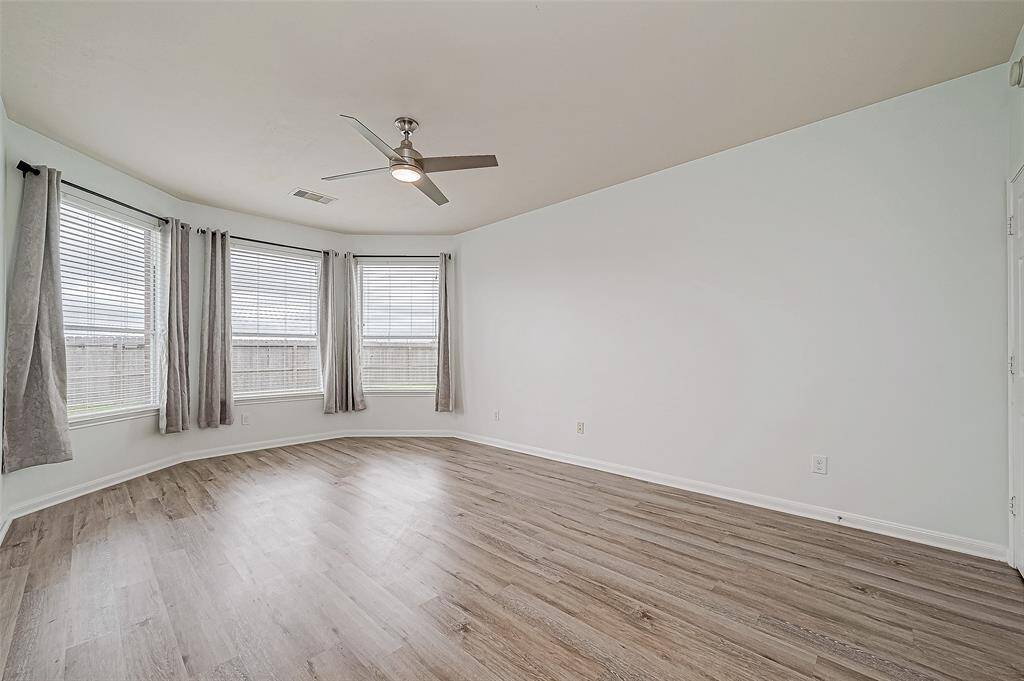 Bright and spacious primary bedroom features a large bay windows with gray curtains, sleek light wood flooring, and a modern ceiling fan.