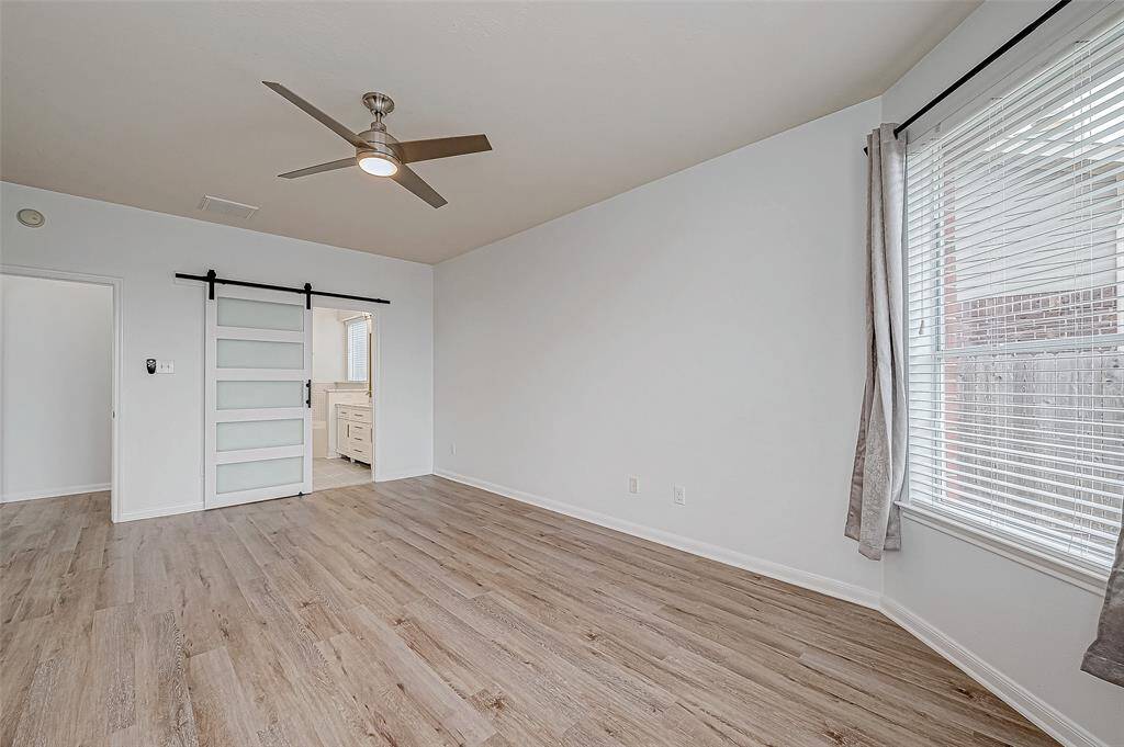 Another view of the primary bedroom designed with light wood flooring, a ceiling fan, and ample natural light from a large window including a stylish sliding barn door leading to an adjacent space, offering a contemporary and open feel.