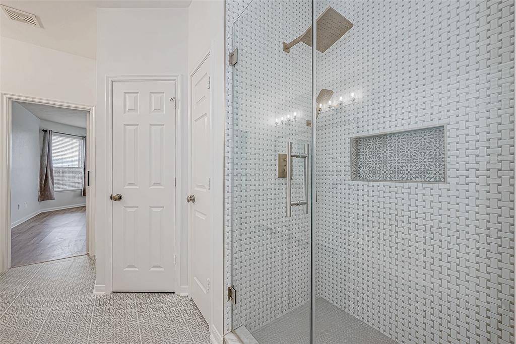 This photo showcases a modern bathroom with a stylish glass-enclosed shower featuring intricate tile work. The space is bright, with light-colored walls and flooring. A doorway leads to a room with a large window, providing ample natural light.