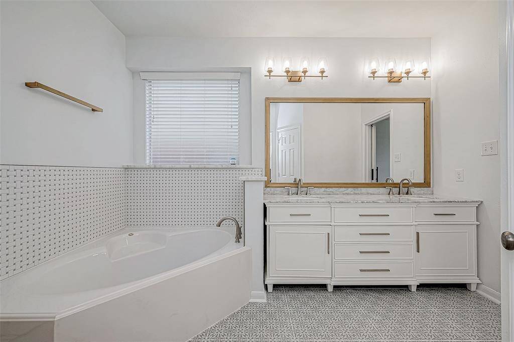 This modern bathroom features a spacious corner bathtub, elegant dual-sink vanity with marble countertop, large mirror, and stylish lighting. The room has a light, airy feel with patterned floor tiles and a window for natural light.