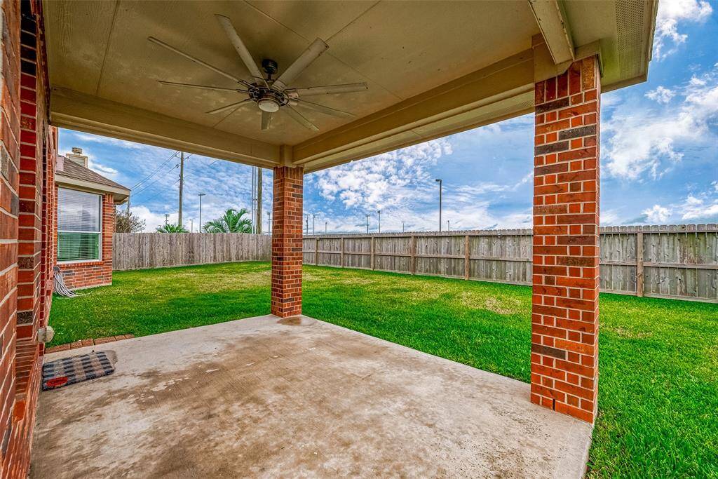 This photo shows a covered patio with a ceiling fan, brick pillars, and a view of a spacious, fenced backyard with lush green grass. Perfect for outdoor relaxation and entertaining.