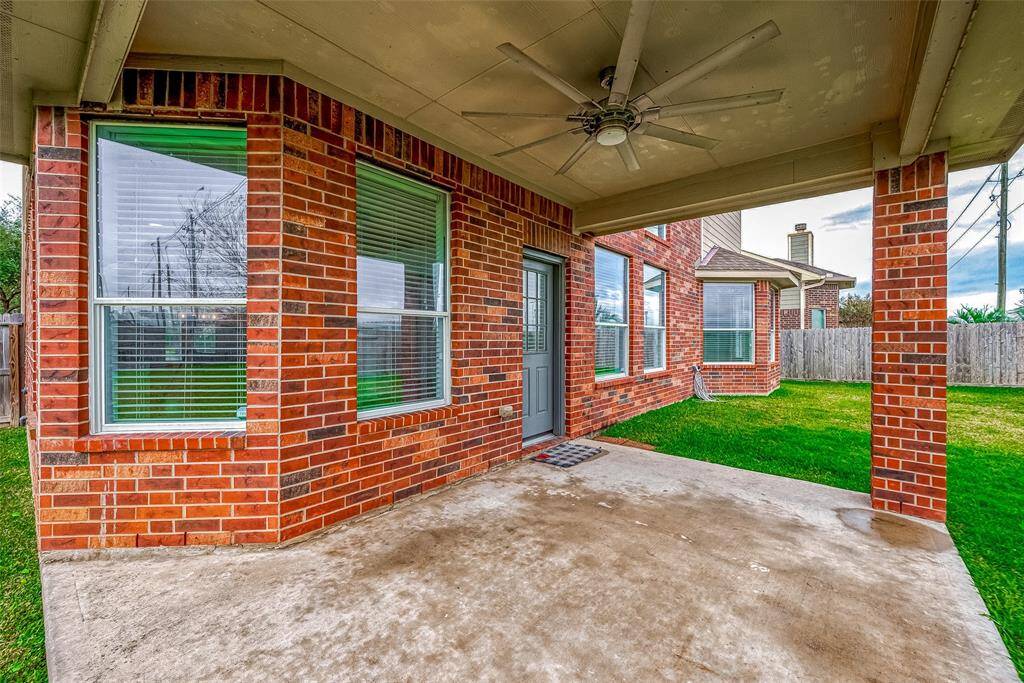 This photo shows a covered patio with a ceiling fan, surrounded by a brick exterior and large windows, overlooking a fenced backyard with a well-maintained lawn.